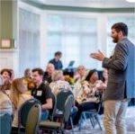 man stands and addresses room of people seated at round tables in Alumni House